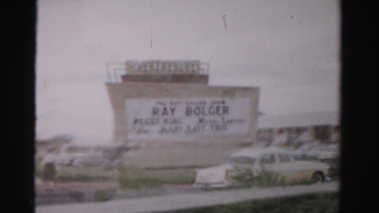 1957 Las Vegas home-movie frame showing the Sahara Hotel sign, marquee lettering, parked cars, and roadside foreground.