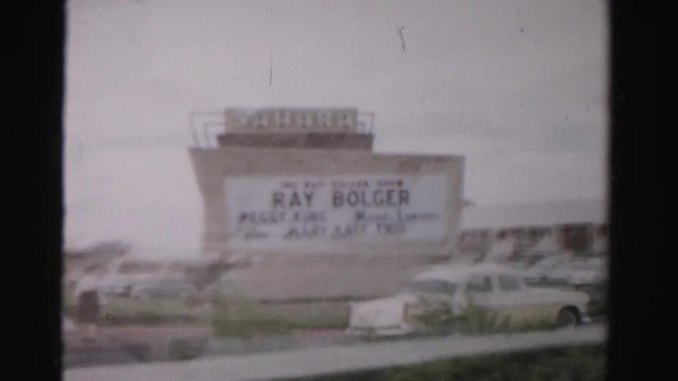 1957 Las Vegas home-movie frame showing the Sahara Hotel sign, marquee lettering, parked cars, and roadside foreground.