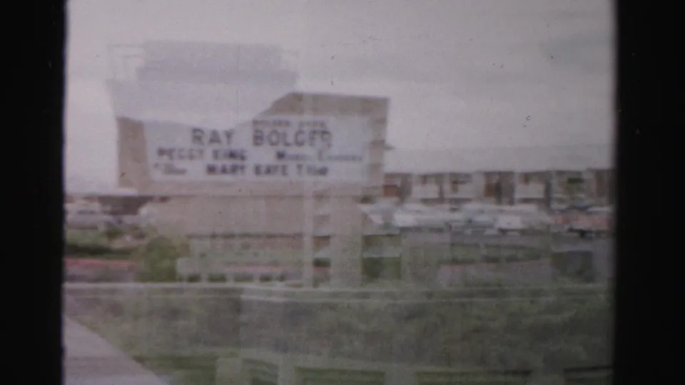1957 Las Vegas 8mm frame showing a closer roadside view of the Sahara marquee, parked cars, landscaping, and low hotel structures.