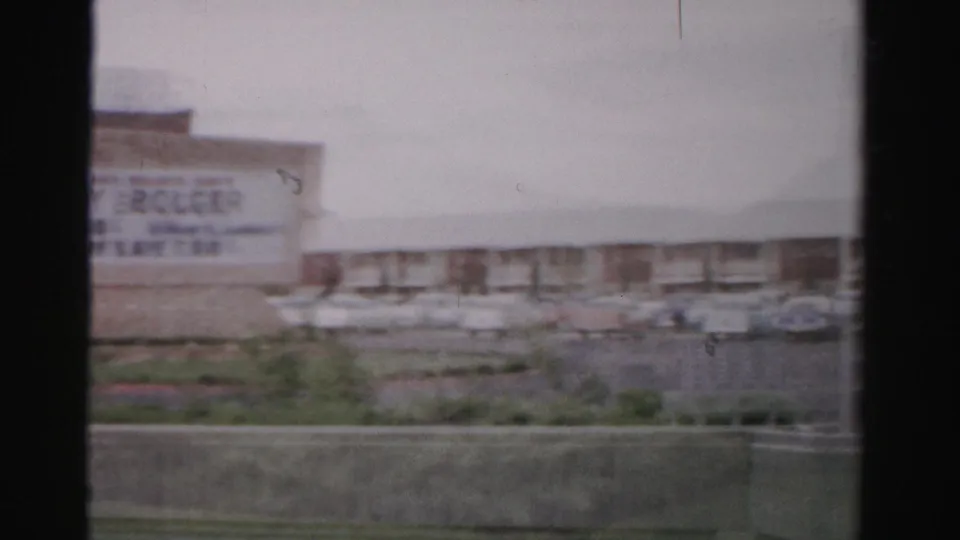 1957 Las Vegas home-movie frame with the Sahara sign partly cropped, cars in the lot, and a low hotel wing behind the roadside scene.