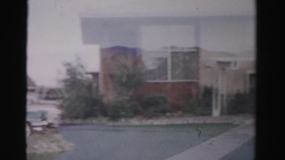 1957 Las Vegas home-movie frame showing motel architecture, a driveway or walkway, landscaping, and cars at the edge of frame.