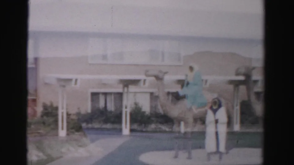 1957 Las Vegas frame showing a standing camel figure or display near a motel entrance, with small indistinct people nearby.
