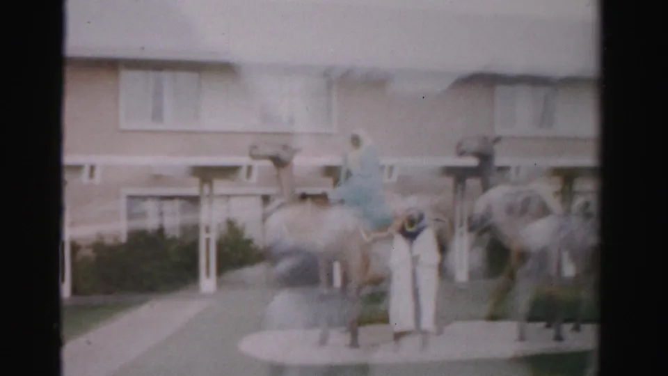 1957 Las Vegas home-movie frame showing camel sculptures, visitors, and costumed figures in front of a beige low-rise building.