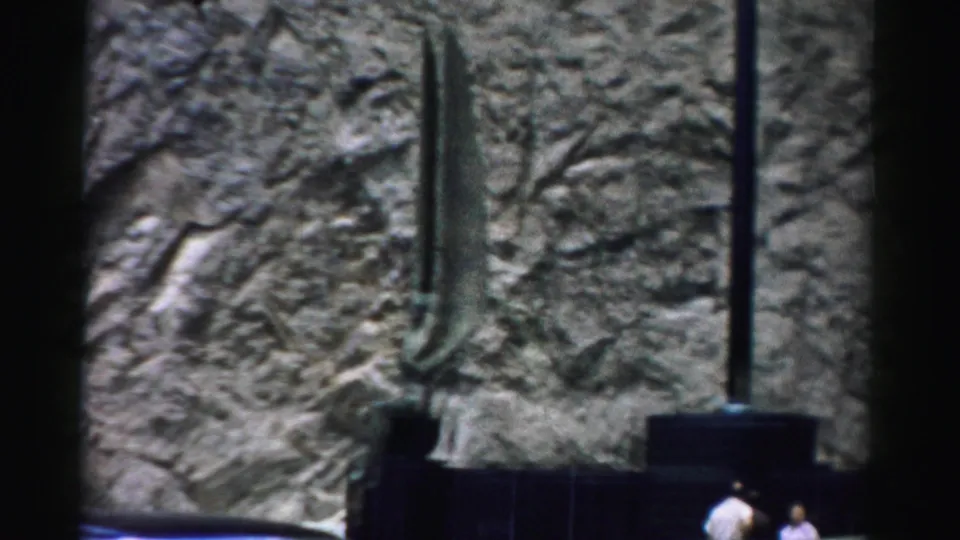 Outdoor sculpture detail with rough wall, metal forms, dark railing, and distant visitors along the lower edge of the 1957 home-movie frame.