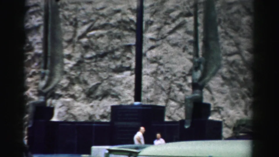 Two visitors observe a large textured outdoor wall sculpture with vertical metal elements in a 1957 Nevada home-movie frame.