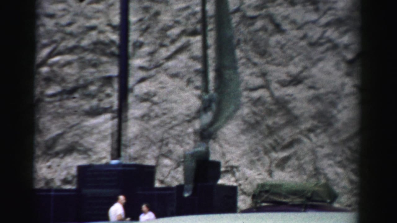 1957 Nevada home-movie frame showing two men below tall green antenna-like forms beside a rugged rock face.