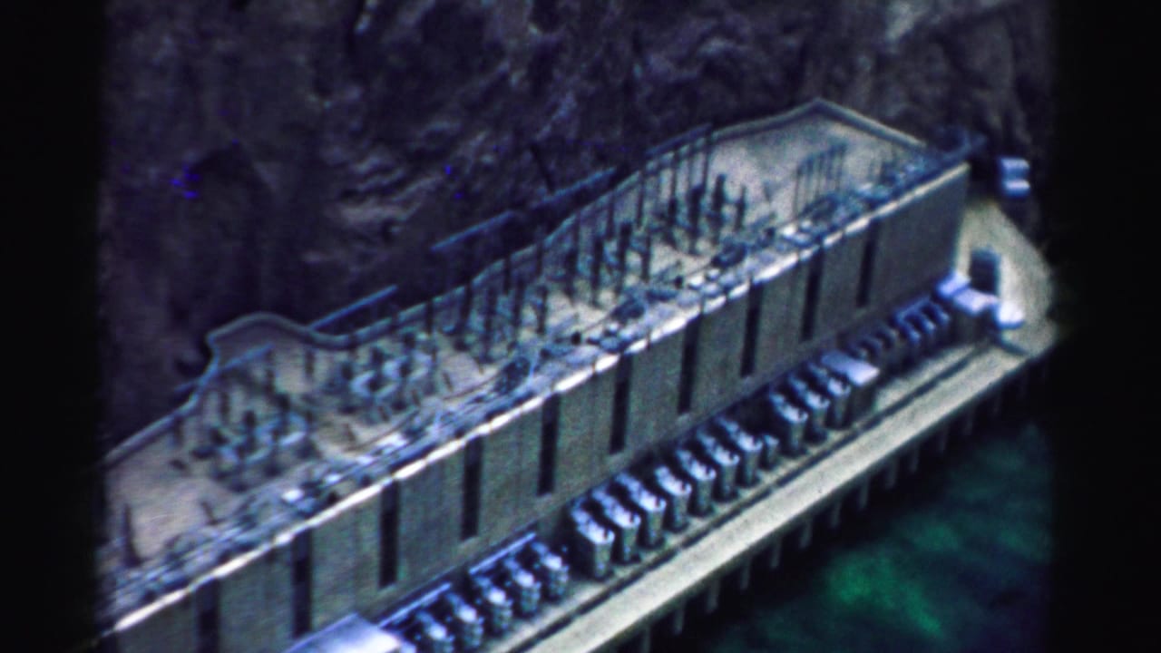 1957 8mm frame showing Hoover Dam infrastructure from above, with canyon walls and blue-green water at the edge.