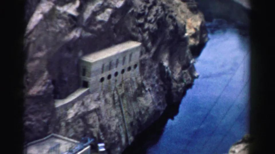1957 8mm view of Hoover Dam from above, showing a concrete structure, cliff face, and deep blue water below.