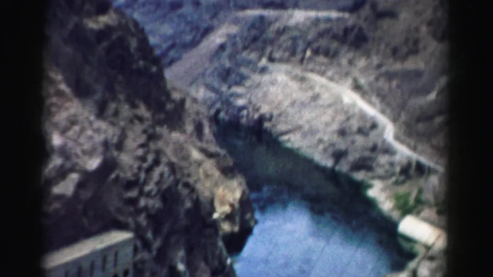 1957 home-movie frame looking across the Hoover Dam canyon setting, with water, rock walls, and paved infrastructure visible.