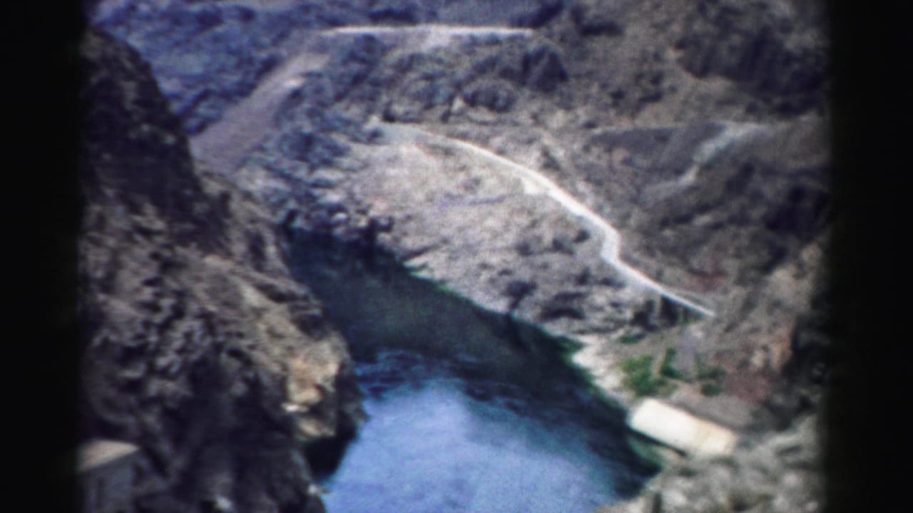 1957 Nevada 8mm frame showing reservoir water, steep canyon rock, and a winding road above a dam area.