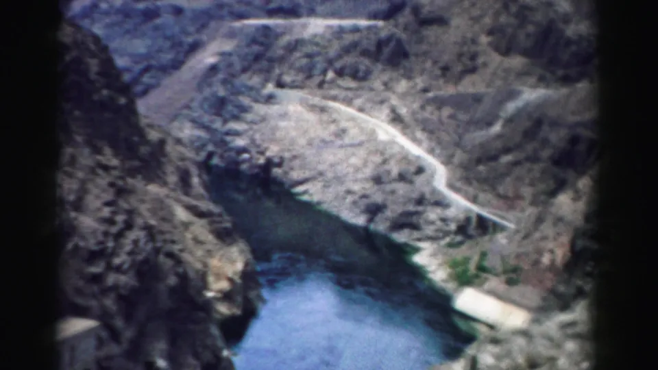 1957 Nevada 8mm frame showing reservoir water, steep canyon rock, and a winding road above a dam area.
