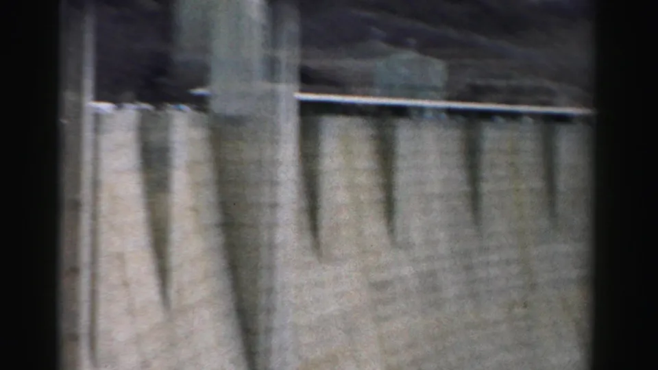 1957 Nevada 8mm frame showing the concrete face of a canyon dam with vertical buttress-like forms and top railing.