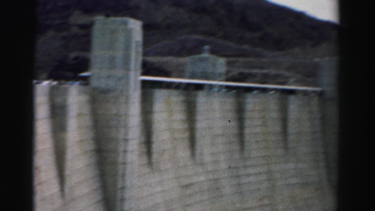 1957 Nevada 8mm frame showing a large concrete dam wall, tower structures, railings, and rugged mountain terrain.