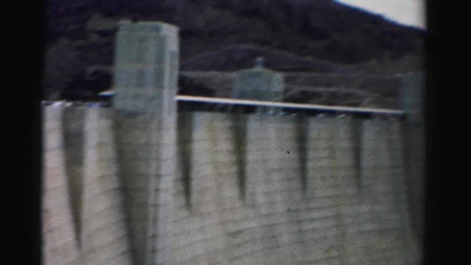 1957 Nevada 8mm frame showing a large concrete dam wall, tower structures, railings, and rugged mountain terrain.