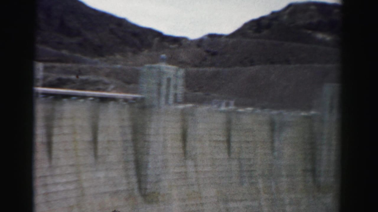 Curved concrete dam face with tower and rocky hills behind it.