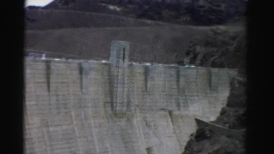 1957 Nevada 8mm frame showing the dam wall from a higher angle, with a central tower and rocky canyon edge nearby.