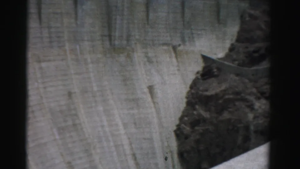 1957 Nevada 8mm frame showing a tall concrete dam face, rocky outcrop, and dark canyon slope with no people visible.