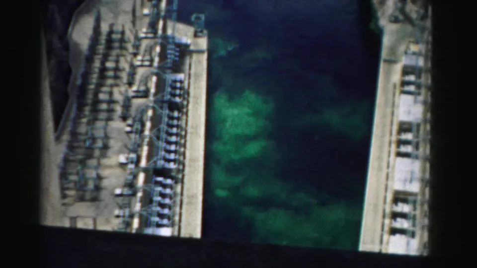1957 Nevada home-movie frame looking down along concrete infrastructure, electrical equipment, and green water in a canyon-like setting.