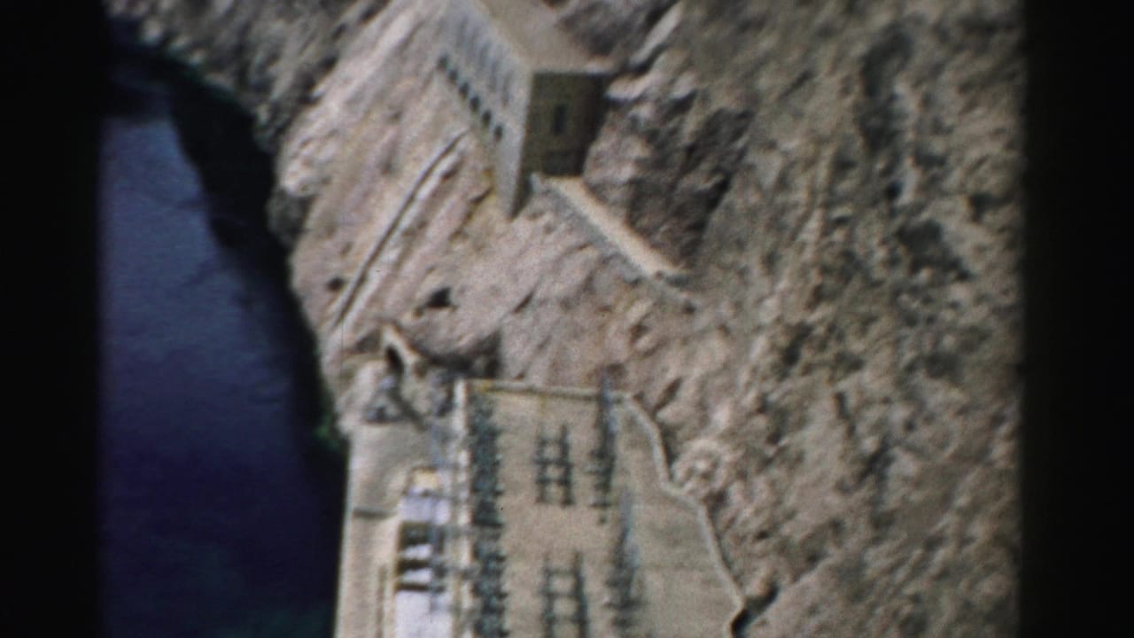 1957 Nevada 8mm frame showing dark river water below rocky cliffs with concrete dam-related buildings and power equipment.