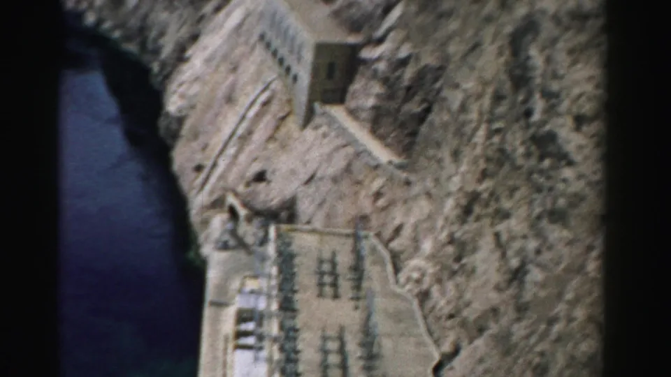 1957 Nevada 8mm frame showing dark river water below rocky cliffs with concrete dam-related buildings and power equipment.