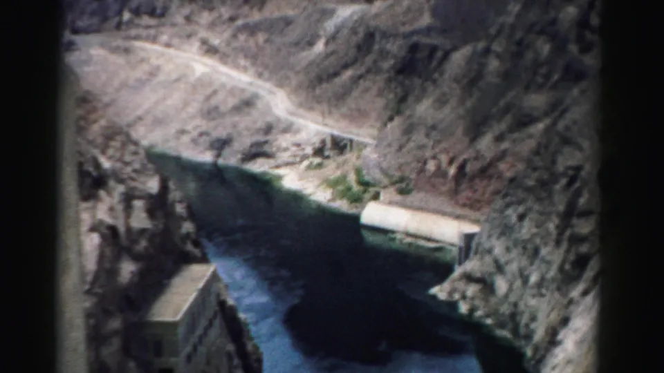 1957 Nevada 8mm view of a canyon river bordered by rocky slopes, a road, and a rectangular concrete facility.
