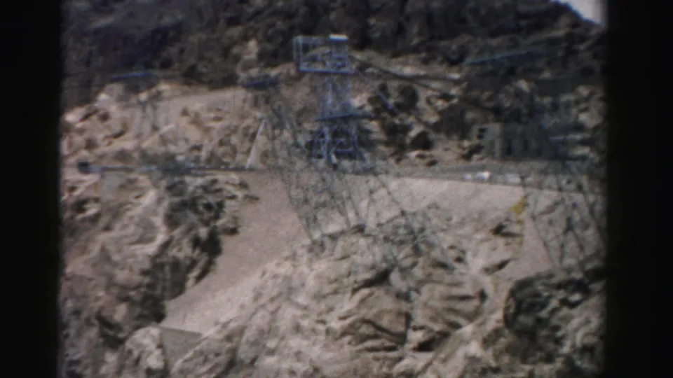1957 Nevada 8mm frame showing transmission towers, power lines, roads, and dam-related structures across a rocky canyon slope.