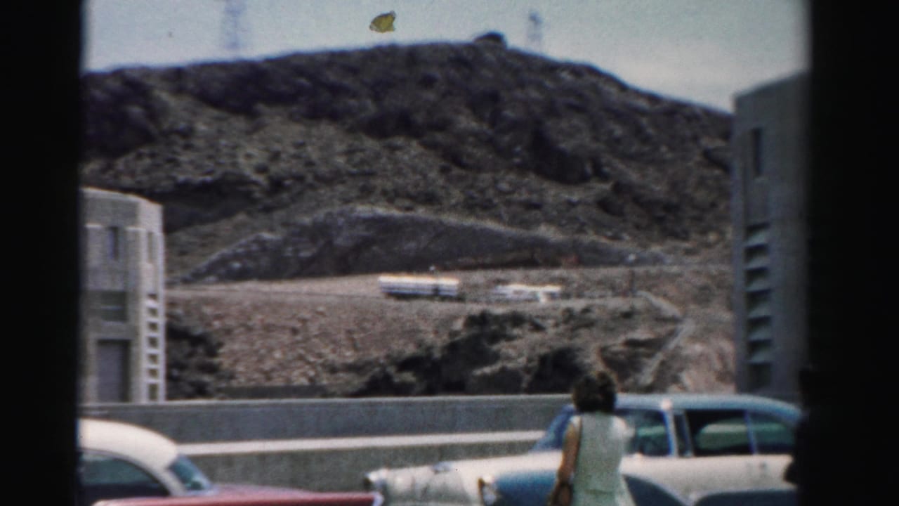Woman near parked classic cars with rocky hill and buildings behind her.