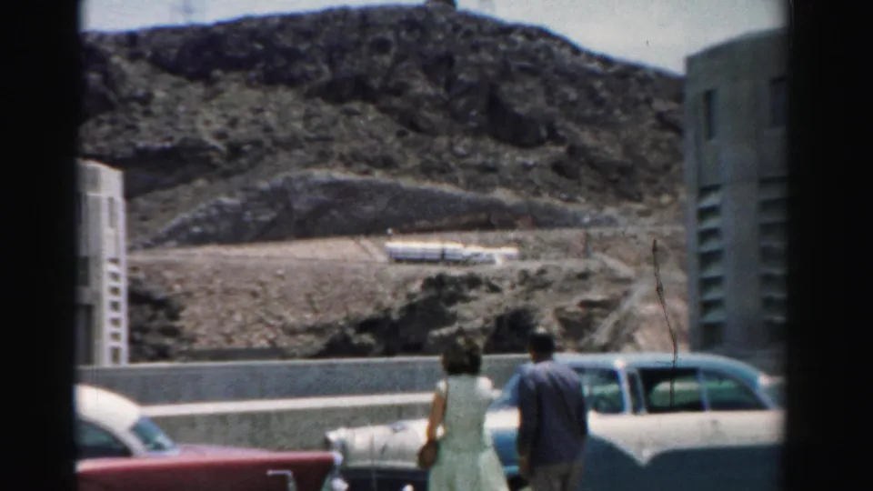 Man and woman walk near classic cars along a roadside barrier in a 1957 Nevada home-movie view of rocky terrain and buildings.