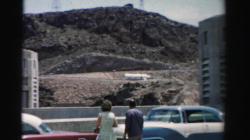 Two adults seen from behind beside parked cars, with a rocky Nevada hillside, roadside wall, and modernist building edge in the background.
