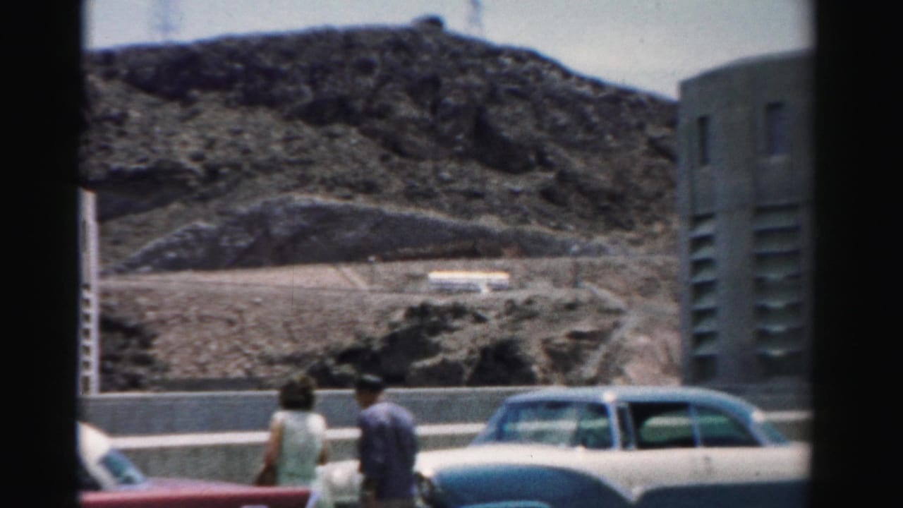 Couple walking beside parked classic cars with rocky hills behind them.