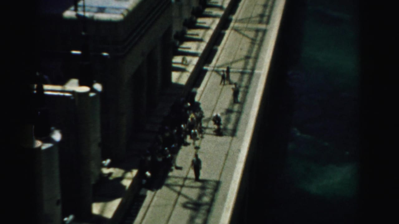 High-angle 1950s Nevada view of tourists moving along an elevated dam walkway beside concrete forms and water.