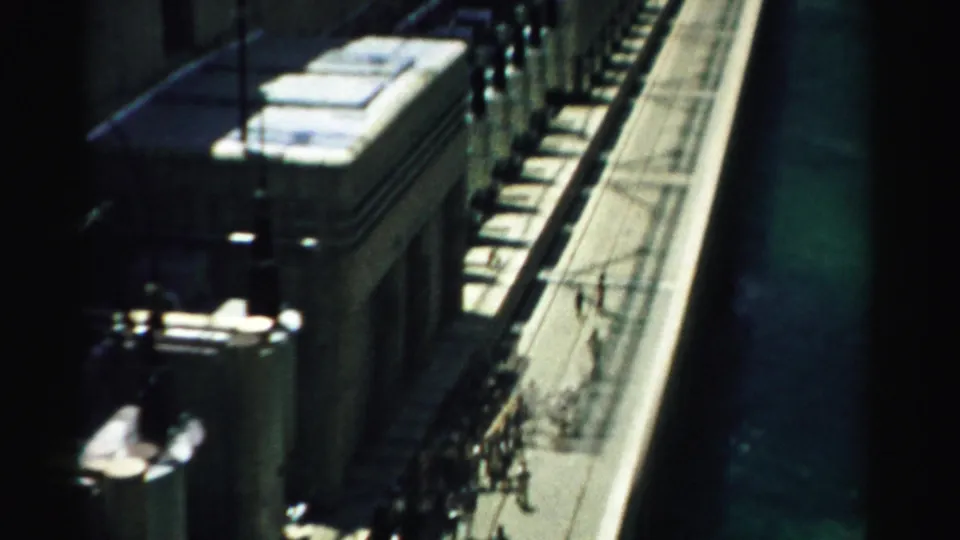 Visitor-shot 1957 Nevada footage looks down along a dam walkway with people, railings, concrete structures, and green water below.