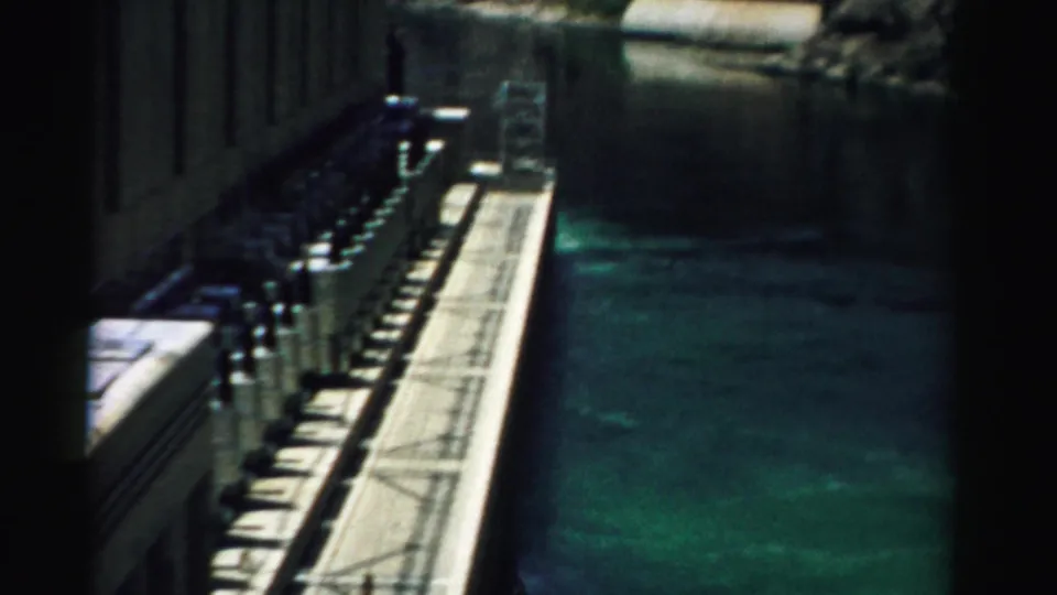 A 1957 Nevada dam view shows a walkway, concrete wall, railings, small visitors, and green water in shadow.