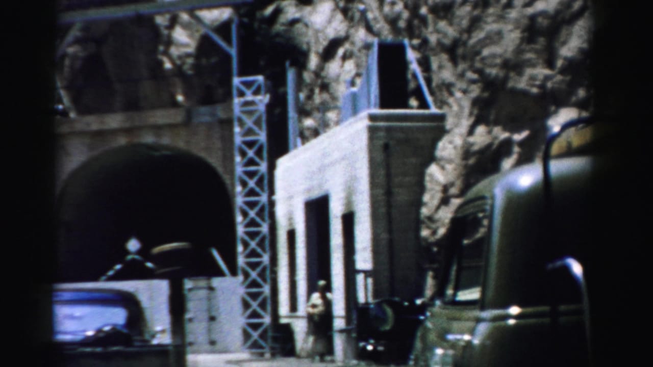 1957 Nevada 8mm frame showing a stone structure embedded in a boulder-like rock face, with metal supports, a vehicle, and a small visitor figure.