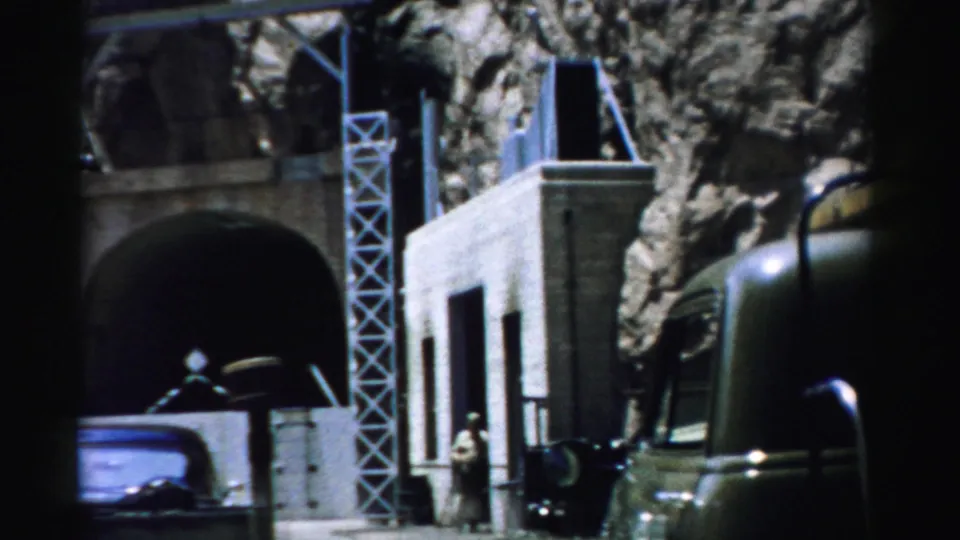 1957 Nevada 8mm frame showing a stone structure embedded in a boulder-like rock face, with metal supports, a vehicle, and a small visitor figure.