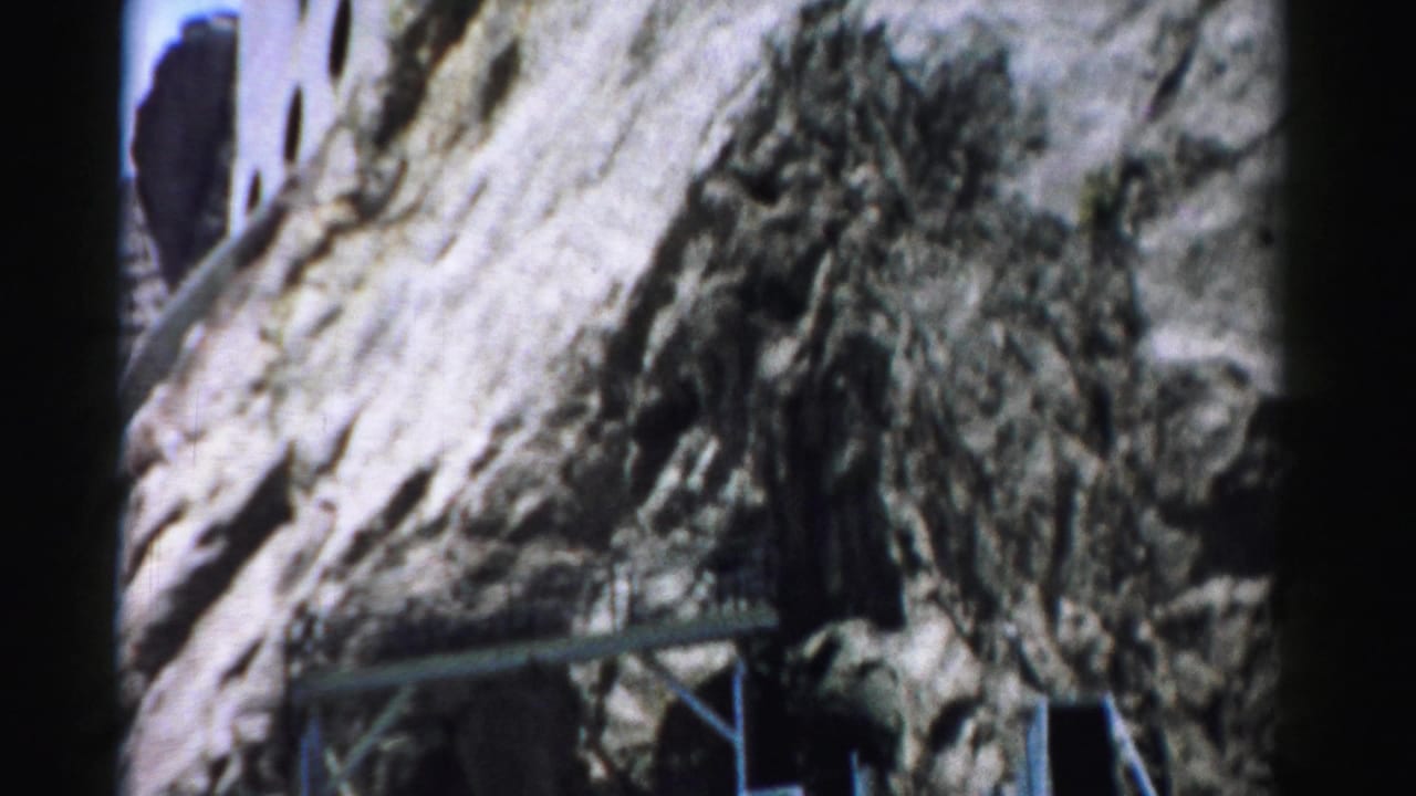 1957 Nevada frame focusing on rough cliff rock beside the embedded structure, with metal bracing and part of the building visible.