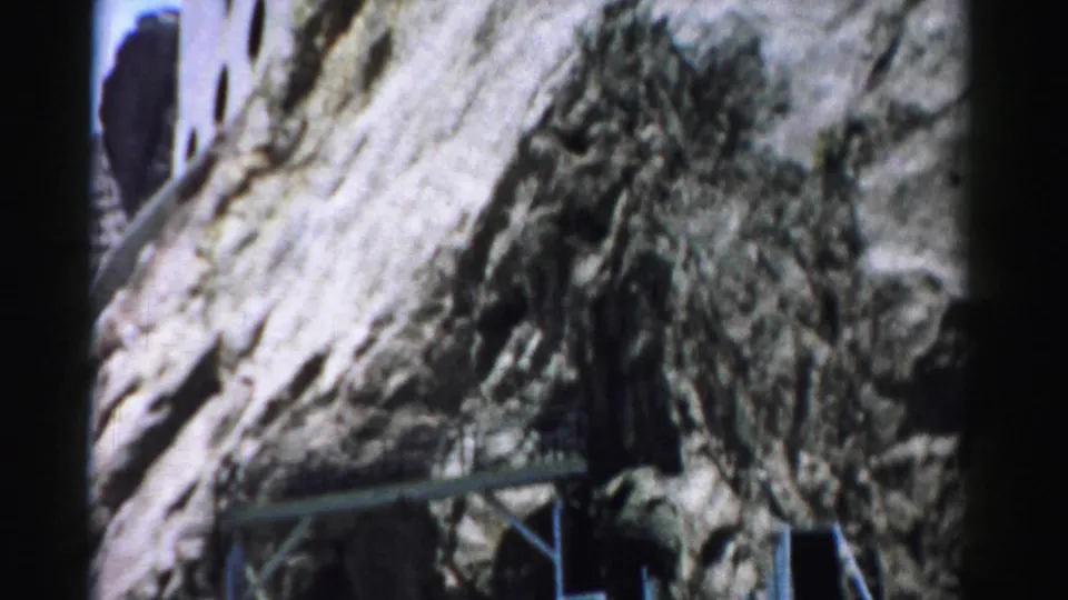 1957 Nevada frame focusing on rough cliff rock beside the embedded structure, with metal bracing and part of the building visible.