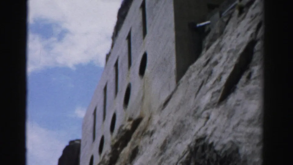 1957 Nevada 8mm frame looking up at the rectangular-windowed stone facade pressed against the rock face under a bright sky.
