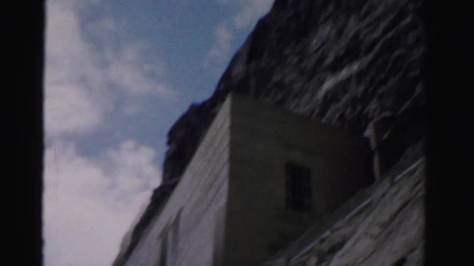 1957 Nevada frame showing part of the embedded stone structure with a dark window, steep rock above, and open sky beyond.