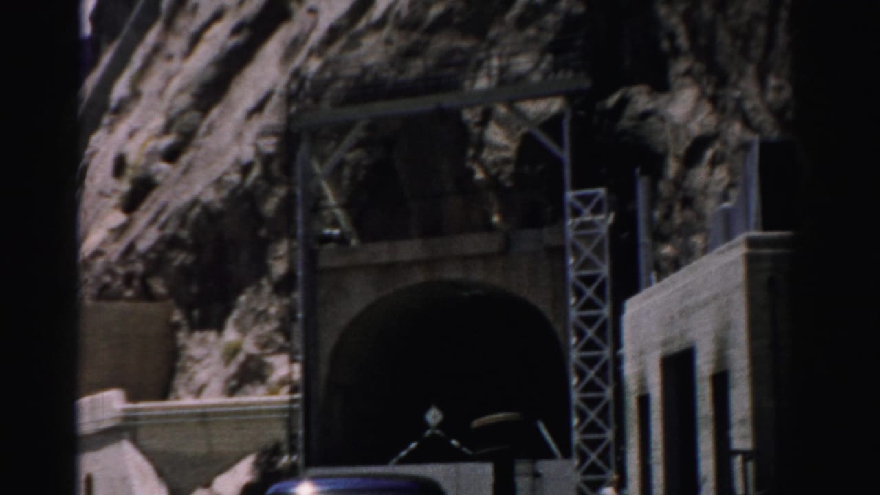 1957 Nevada 8mm frame showing rocky cliffs around a dark tunnel portal, concrete retaining walls, metal scaffolding, and a partial vehicle.