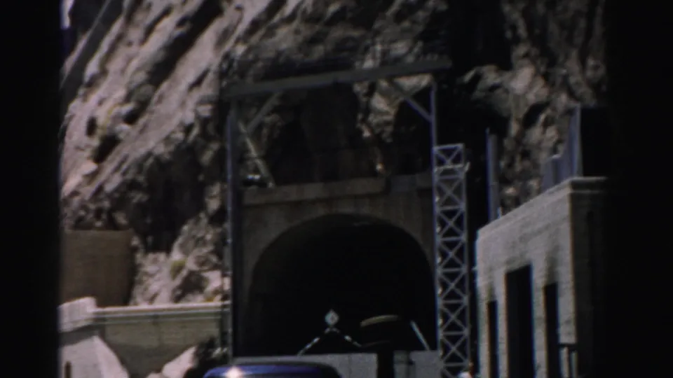1957 Nevada 8mm frame showing rocky cliffs around a dark tunnel portal, concrete retaining walls, metal scaffolding, and a partial vehicle.