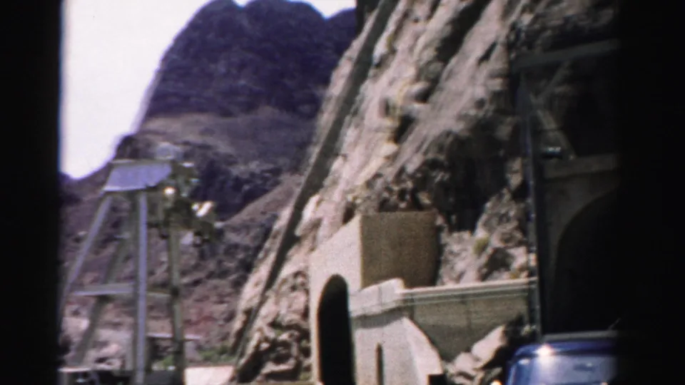 1957 Nevada 8mm frame looking past concrete tunnel structures toward a metal shaft structure on a rocky desert mountain slope.