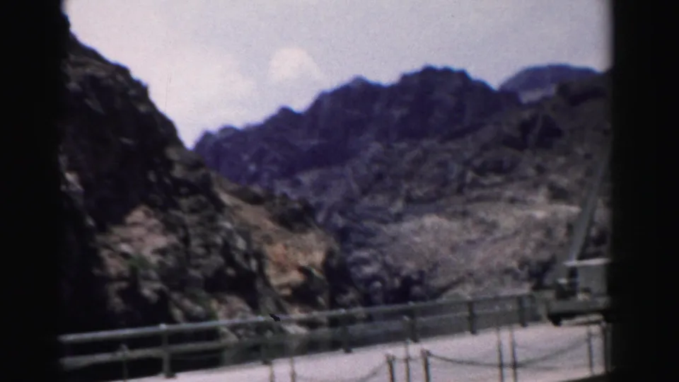 1957 Nevada 8mm frame of roadside railing and concrete infrastructure below rugged rocky mountains in an arid landscape.