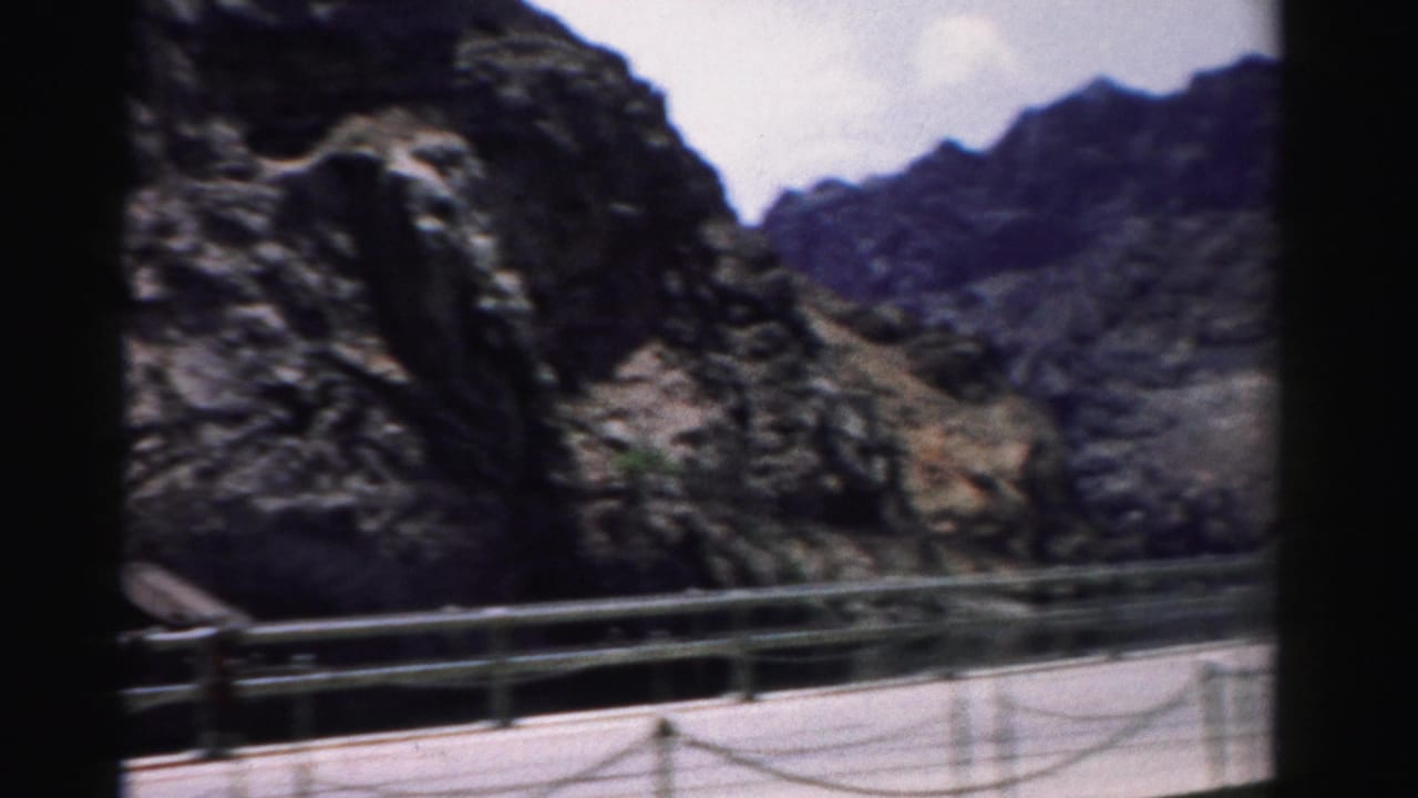 1957 Nevada home-movie frame showing a paved mountain walkway bordered by metal rails beneath steep rock faces.