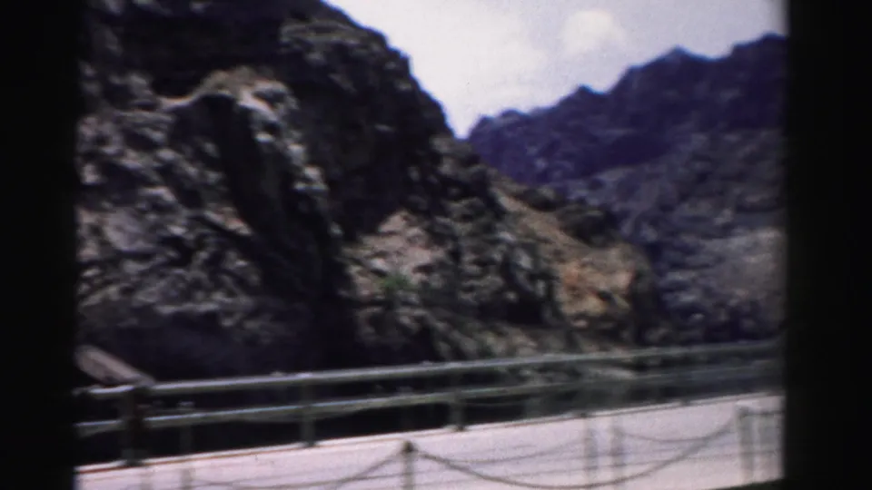 1957 Nevada home-movie frame showing a paved mountain walkway bordered by metal rails beneath steep rock faces.