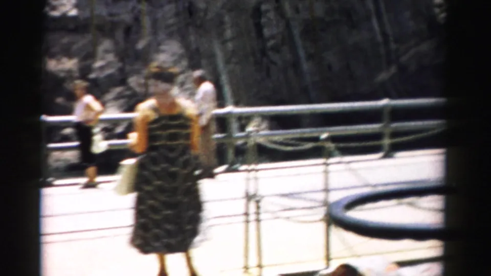Visitors in 1950s summer attire move along a paved Nevada overlook beside rope barriers and metal guardrails.