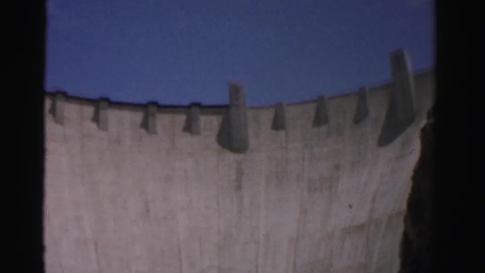 Low-angle 1957 8mm frame of Hoover Dam's curved upper edge and buttresses against blue sky in Nevada.