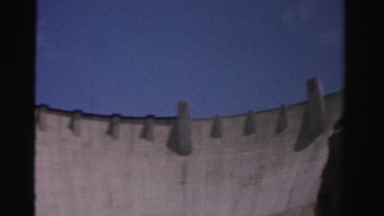 Curved concrete dam wall under a deep blue sky, framed by dark film edges.