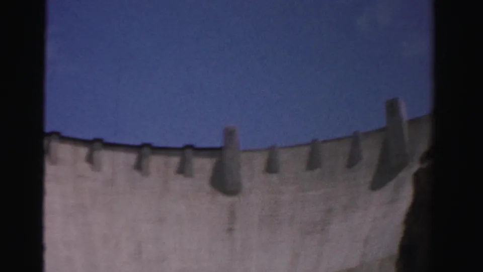 1957 8mm view of Hoover Dam in Nevada, showing a curved concrete wall against blue sky with soft focus and dark frame edges.
