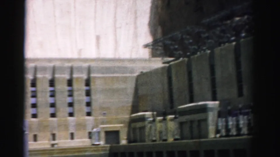 1957 8mm frame of Hoover Dam, with bright concrete face, adjacent tower structure, vertical openings, and dark film-edge shadow.